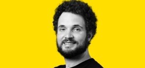 Fundo amarelo, retrato de um homem em branco e preto por volta dos 30 anos, com barba e cabelo. Ele sorri para a foto