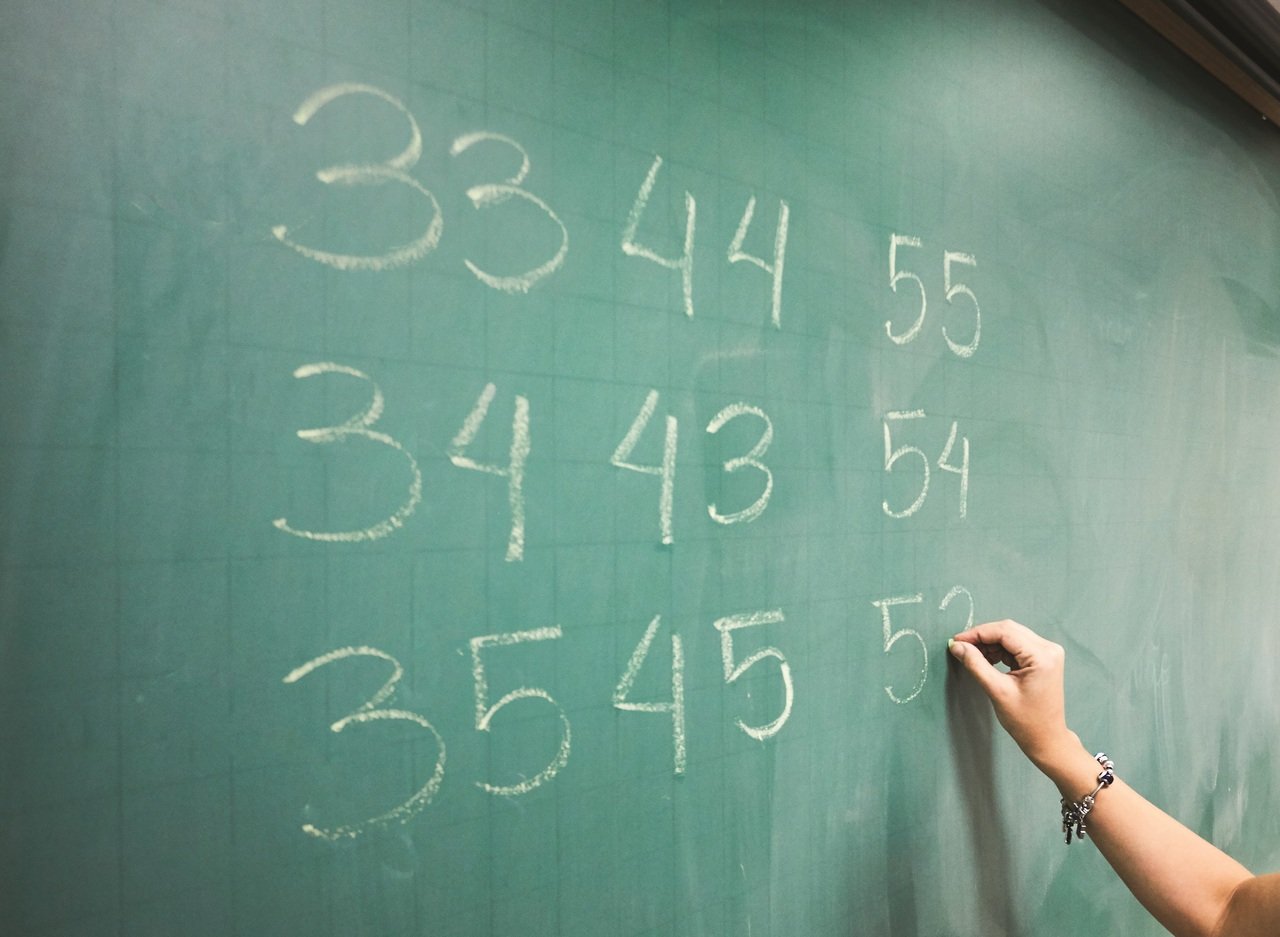 Detalhe de foto na qual a professora escreve n&uacute;meros com giz branco em uma lousa verde