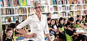 Retrato da professora Selene Coletti. Ao fundo, vê-se uma sala de aula de crianças de cerca de 7 anos.