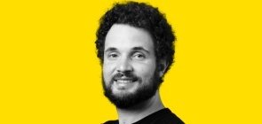 Fundo amarelo, retrato de um homem em branco e preto por volta dos 30 anos, com barba e cabelo. Ele sorri para a foto