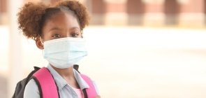 Uma menina negra de cerca de 7 anos usando máscara contra covid-19 e carregando uma mochila nas costas