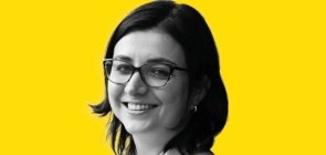 Mulher de óculos, está de lado e sorri para a foto. Ela tem cabelo na altura do ombro