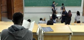 Jovens em uma sala de aula com várias cadeiras vazias e uma lousa com várias inscrições, um jovem negro é visto de costas em primeiro plano